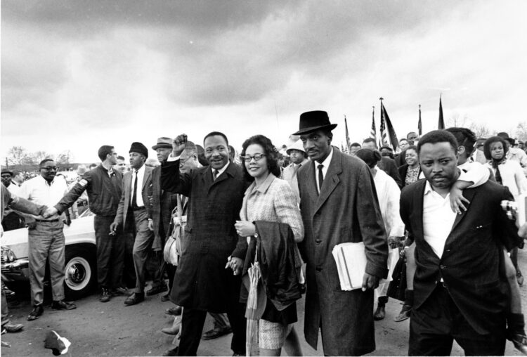 A group of people stand in front of a crowd. Some people are holding U.S. flags up.